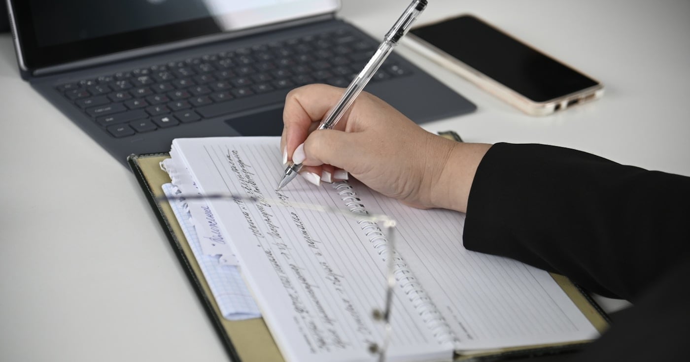 Person writing in a notebook with a laptop in the background