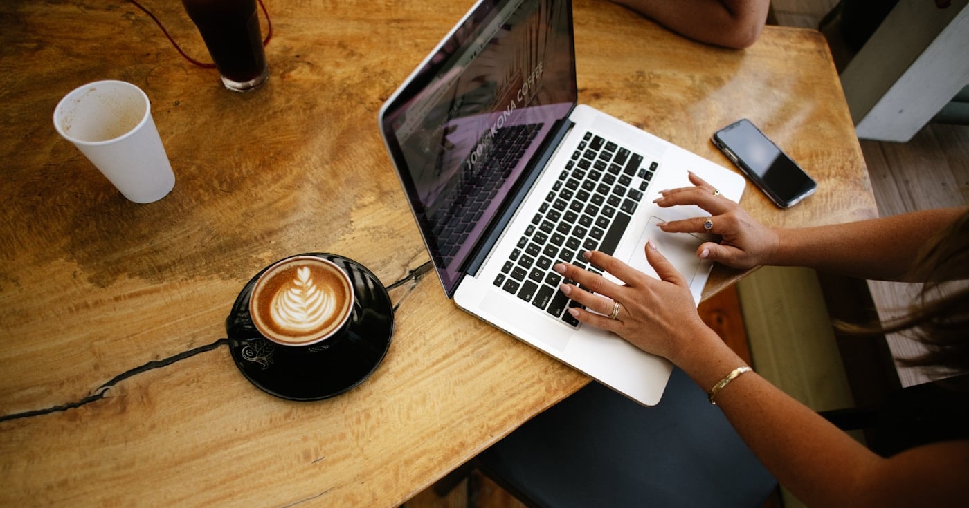 Person working on a laptop at a café table with a coffee, representing freelance article writing