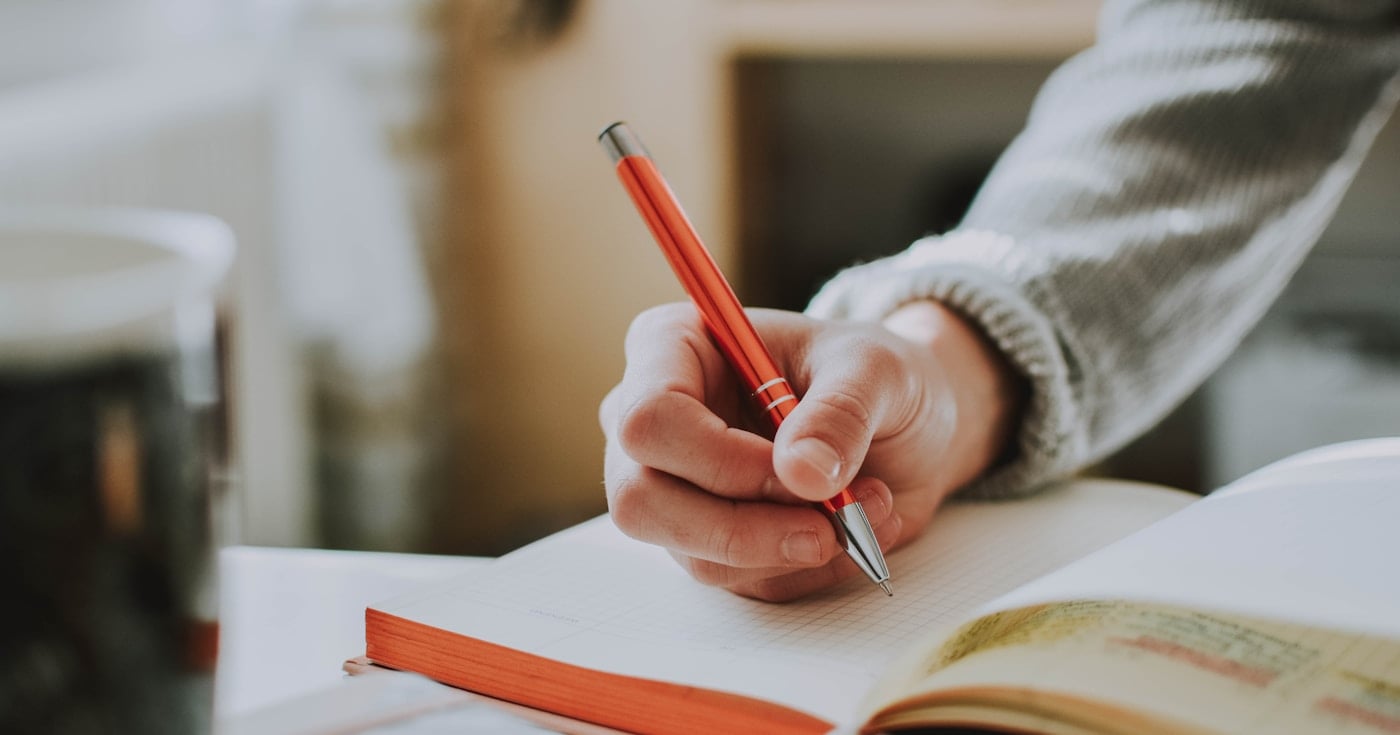 Person writing in notebook with a red pen