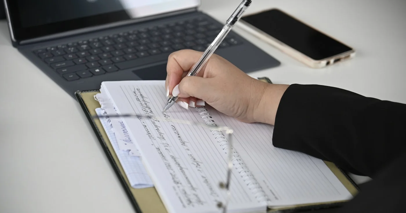 Person writing in a notebook with a laptop in the background