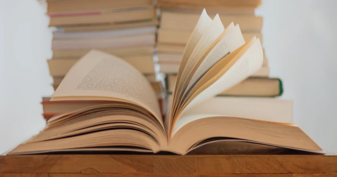 Open book on a wooden table with stacks of books behind it